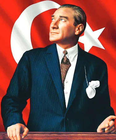 Mustafa Kemal Atatürk ellerini kürsüye dayamış, yukarıya bakıyor; arkasında Türk bayrağı